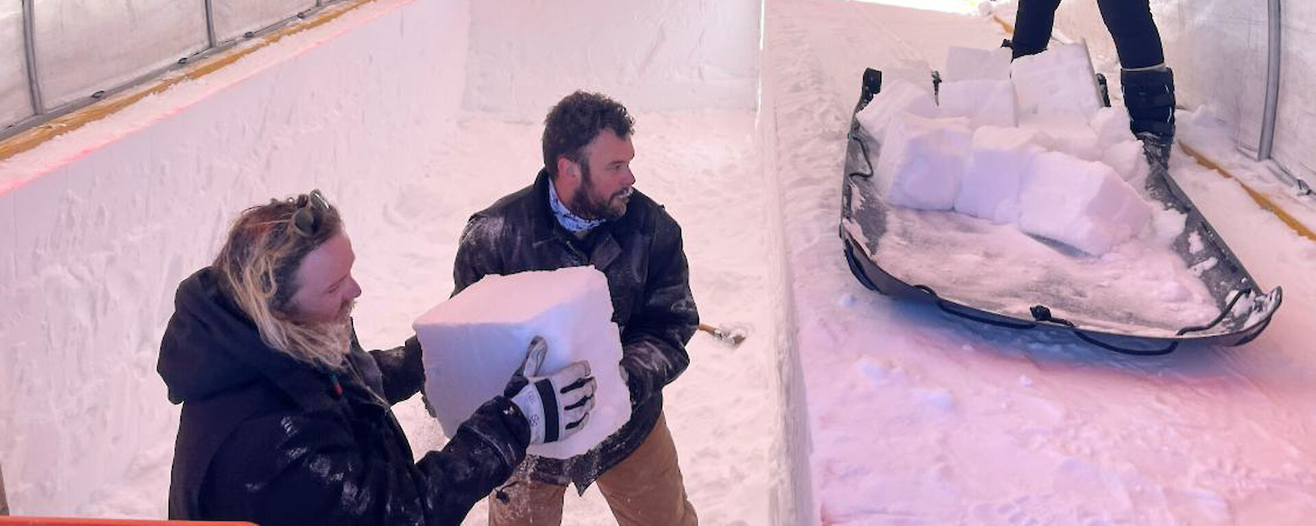 Two men lift a square block of ice out of a trench in the snow towards a woman standing beside a sled loaded with other blocks of ice.