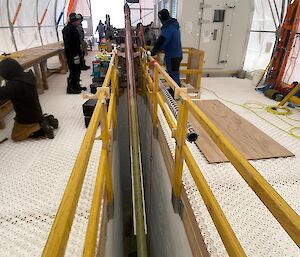A long drill barrel is tilted from horizontal towards verticle inside a 6 metre deep channel cut in the ice.