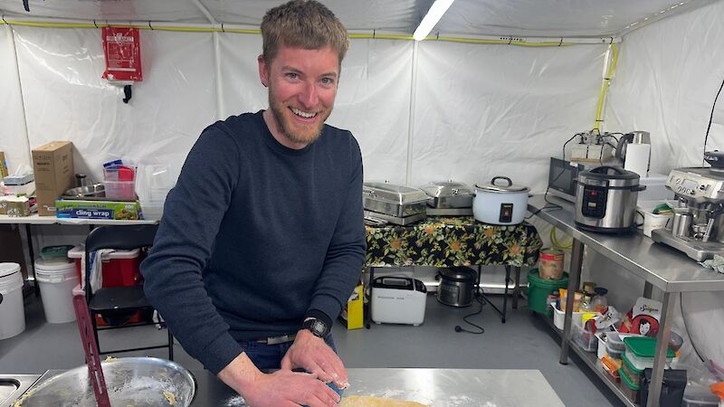 A man uses a wter bottle to roll a small circle of dough on a steel benchtop dusted with flower.