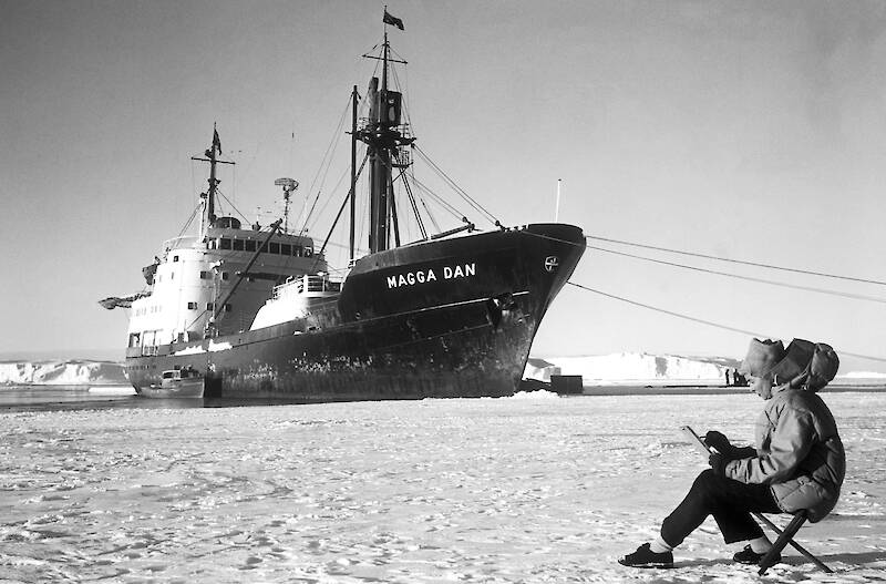 A woman sitting on a camp chair on ice, with the Antarctic ship Magga Dan anchored in a bay about 100 metres away.
