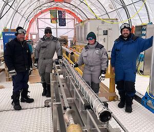 Four people standing beside a cylindrical metal core barrel, lying horizontally across a metal frame.