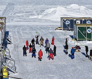 Aerial view of people playing a game of volleyball in between two rows of shipping containers, with a pile of snow in the background.