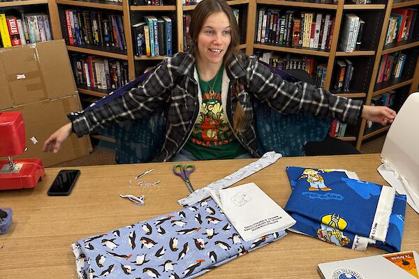 Lisa the Fix it Fairy at the library desk with her sewing machine and donning her fairy wings in the foreground, the library shelves in the background.