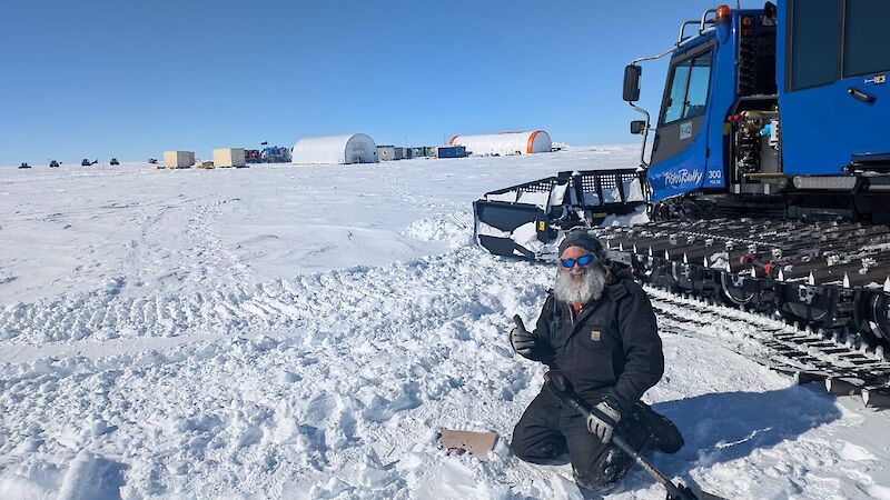 A man kneeling in snow with a shovel. A small square lid is visible beside him, which sits above a 10 metre-long temperature string buried in the snow, used to measure surface temperatures.