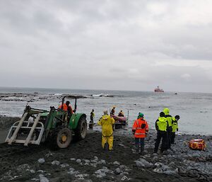 A group of people stand on the sea-shore while a red IRB launches off a boat trailer into the sea.