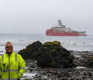 A smiling man in a yellow jacket stands on the sea-shore with a red ship around 500m offshore.