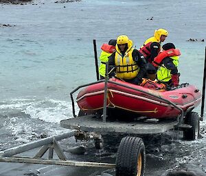 Two men wearing yellow life jackets and helmets crew a red IRB being driven onto a trailer at the water's edge. There are two passengers wearing bright yellow jackets.