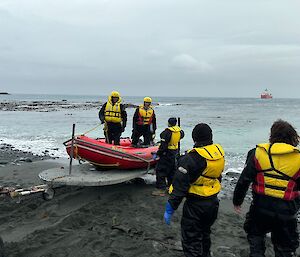 Two men wearing yellow life jackets and helmets standing up in a red IRB loaded onto a trailer at the water's edge.