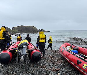 Five men in dry suits and yellow life jackets stand next to two red inflatable rescue boats on the sea-shore. They are all looking out to sea.