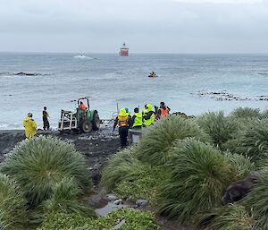 A group of people in bright yellow jackets stand on the sea-shore. A tractor and boat trailer are at the water's edge, and IRB is offshore and a large red ship is around 500m offshore.