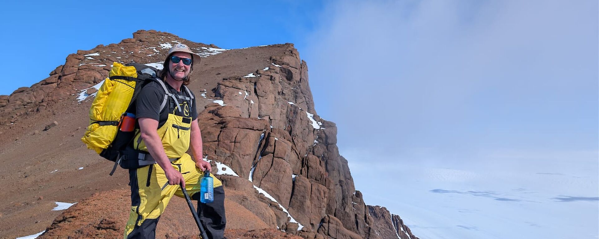 A person stands on a brown rocky slope with ice behind him and blue sky above