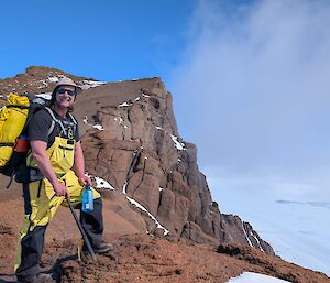 A person stands on a brown rocky slope with ice behind him and blue sky above