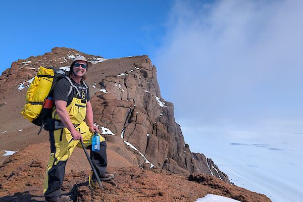 A person stands on a brown rocky slope with ice behind him and blue sky above