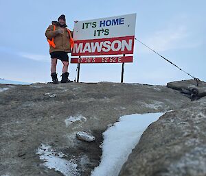 A person stands on a rock, next to a sign saying 'It's home, it's Mawson'