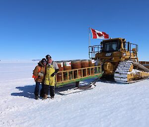 Two people stand near a dozer on a snowy slope