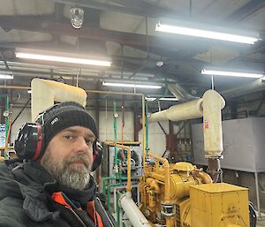 A man wearing a black jacket and hearing protection stands in front of a large yellow generator