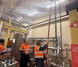 two people in high vis extend a long red pole towards the ceiling of a workshop area