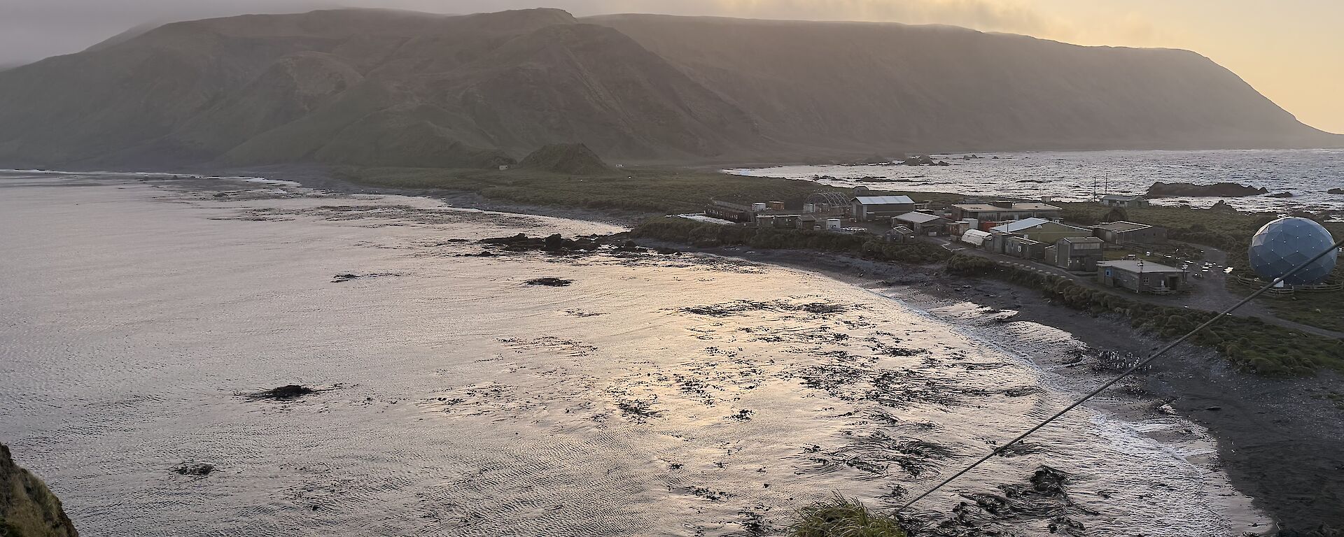 The sun sets over a distant plateau of an island. There is a narrow isthmus in the mid-ground joining a hill in the foreground.