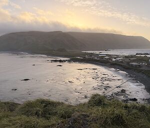 The sun sets over a distant plateau of an island. There is a narrow isthmus in the mid-ground joining a hill in the foreground.