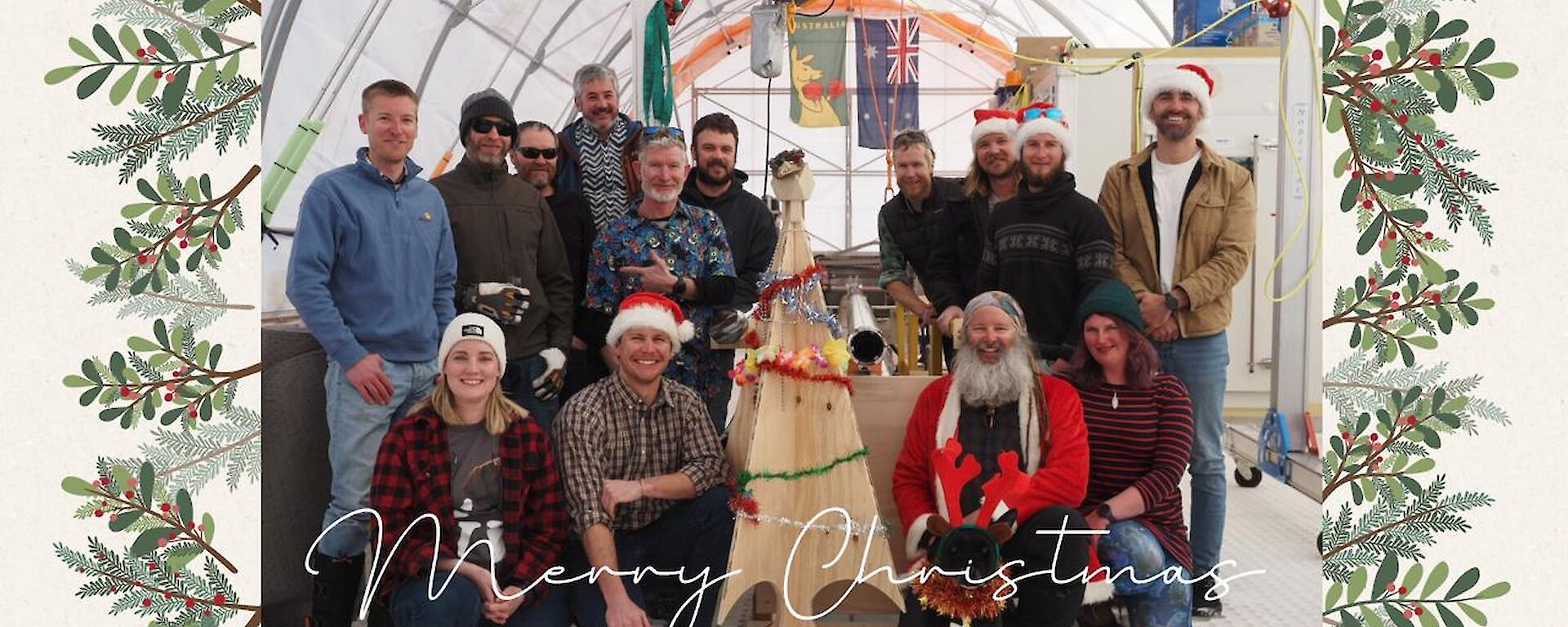 A small group of people, some wearing Santa hats, gathered around a wooden Christmas tree with tinsel inside the drilling shelter for the million year ice core.