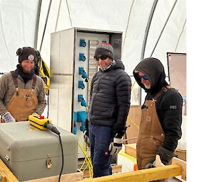 Three men standing beside a metal box that contains controls for a winch.