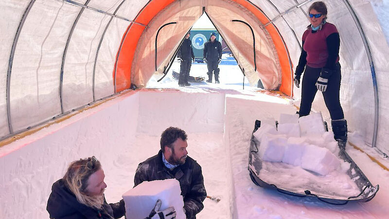 Two men lift a square block of ice out of a trench in the snow towards a woman standing beside a sled loaded with other blocks of ice.