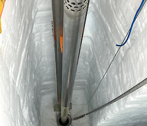 View down into a trench cut in ice, with the metal barrel of an ice core drill hovering above a drill hole.