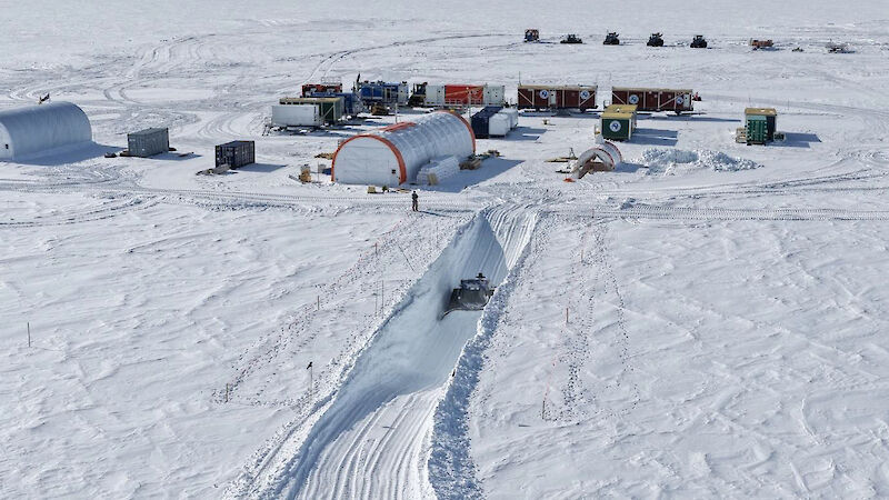 Drone view of a snow groomer deep in a trench cut in the ice, near an ice drilling camp site made up of large cylindrical tents and shipping containers.