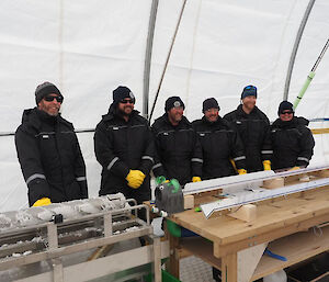 Six people all wearing black drilling suits standing triumphantly beside the first metre of ice drilled this season.