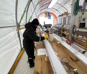 A woman uses a vacuum to remove excess drilling fluid from an ice core positioned in a horizontal metal V-shaped channel on a bench.