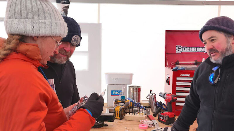 A woman shows a drill cutter to two men, standing at a work bench.