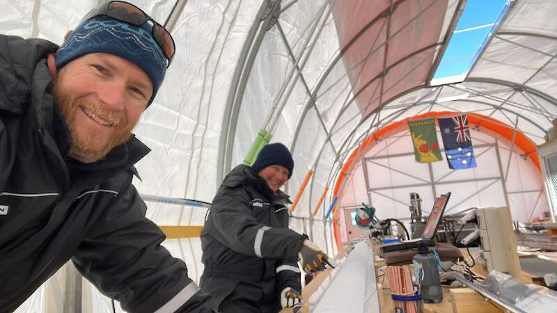 Two people smiling at the camera beside an ice core resting in a metal tray on a bench.