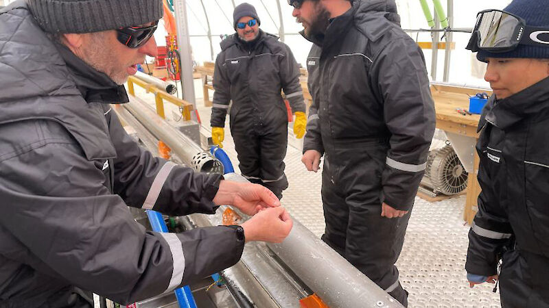 Four people standing beside a horizontal ice drilling barrel. One man is showing a women some broken pieces of metal.