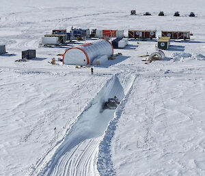 Aerial view of the inland station of tents and shipping containers. A snow groomer is excavating snow inside a deep trench in front of the station.