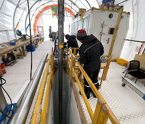 One person uses their hands to guide the vertical barrel of a deep ice core drill into a deep slot in the ice. A second person stands beside some drill controls.