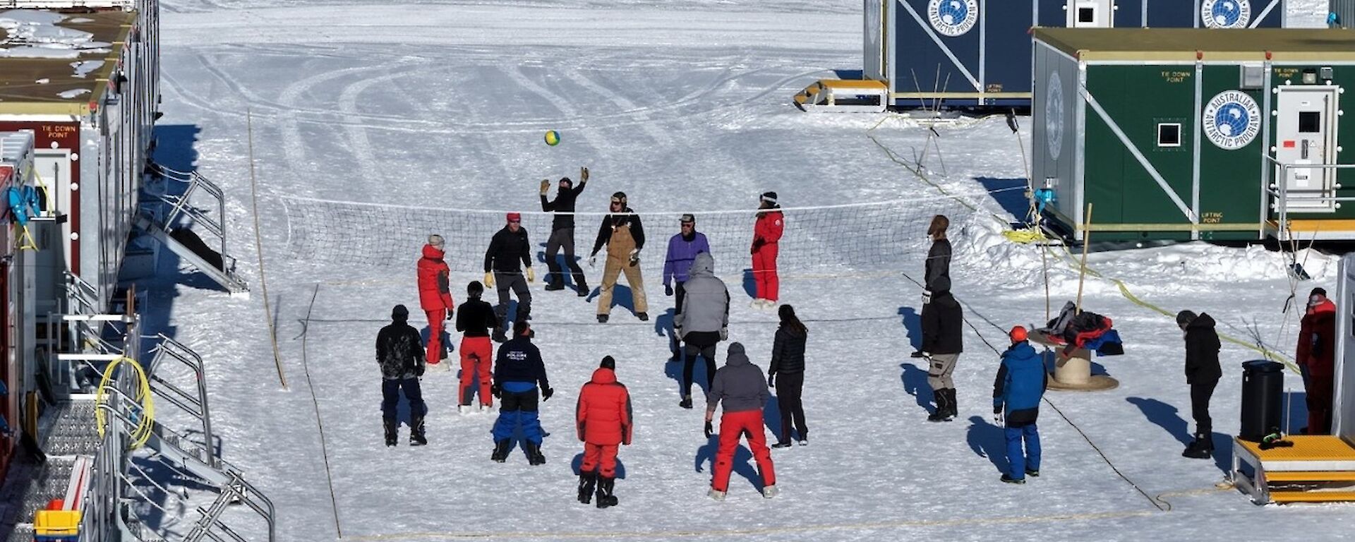 A team of people play volley ball on some flat ice in between shipping containers that make up the accommodation and living areas of the inland station..