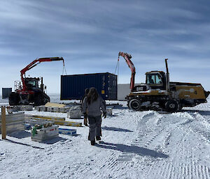 Two tractors use their cranes to lift a shipping container on to a sled.