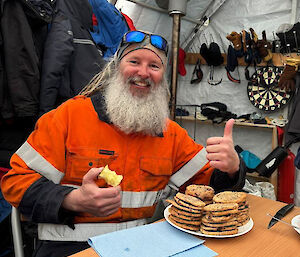 A man holding an apple core in one hand and giving a thumbs up to a large plate of chocolate chip biscuits on a table in front of him.