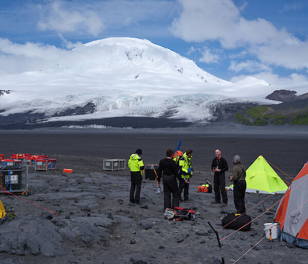 A group of people stand on a black volcanic field with a snow covered volcano behind them and bright yellow and orange tents around