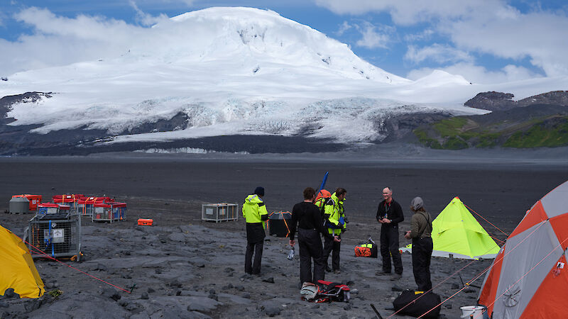 A group of people stand on a black volcanic field with a snow covered volcano behind them and bright yellow and orange tents around
