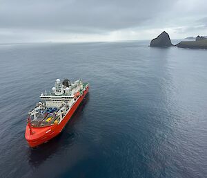 A large red and white ship sits on a glassy grey sea with cloud above and large rock formations in the background