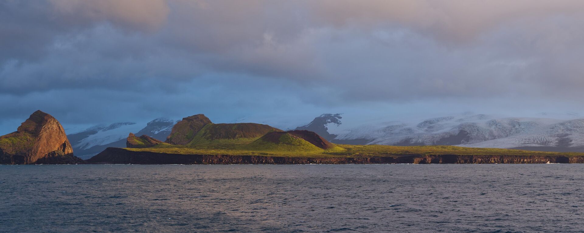 Cloud over dark rock cliffs with a large rock formation to the left and snow covered mountains behind