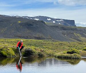 A person in red bends over and takes water from a lake with a white tube