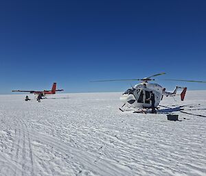 A helicopter and a small plane sit on a snowy plain under a very blue sky