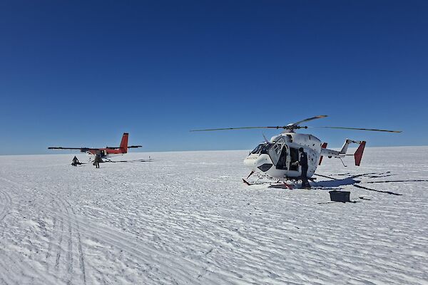 A helicopter and a small plane sit on a snowy plain under a very blue sky