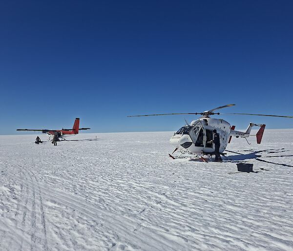 A helicopter and a small plane sit on a snowy plain under a very blue sky
