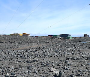A metallic mast is situated in a rocky landscape with yellow and green buildings in the background, under a blue sky.