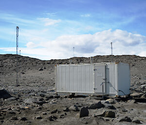 A beige coloured hut and a number of masts in a rocky landscape under a blue sky