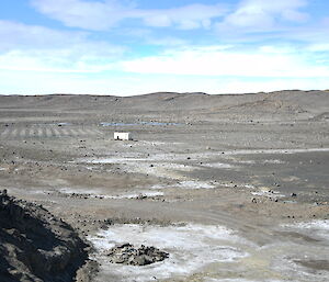 A very wide shot showing a rocky landscape unger a blue sky with some puffy clouds. In the bottom of the valley is a beige hut and an array of metal poles, lined up like a vineyard.