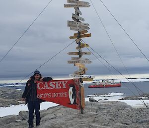 'Turtle' De Meo standing next to the Casey station sign, with the resupply vessel RSV Nuyina pictured in the background.